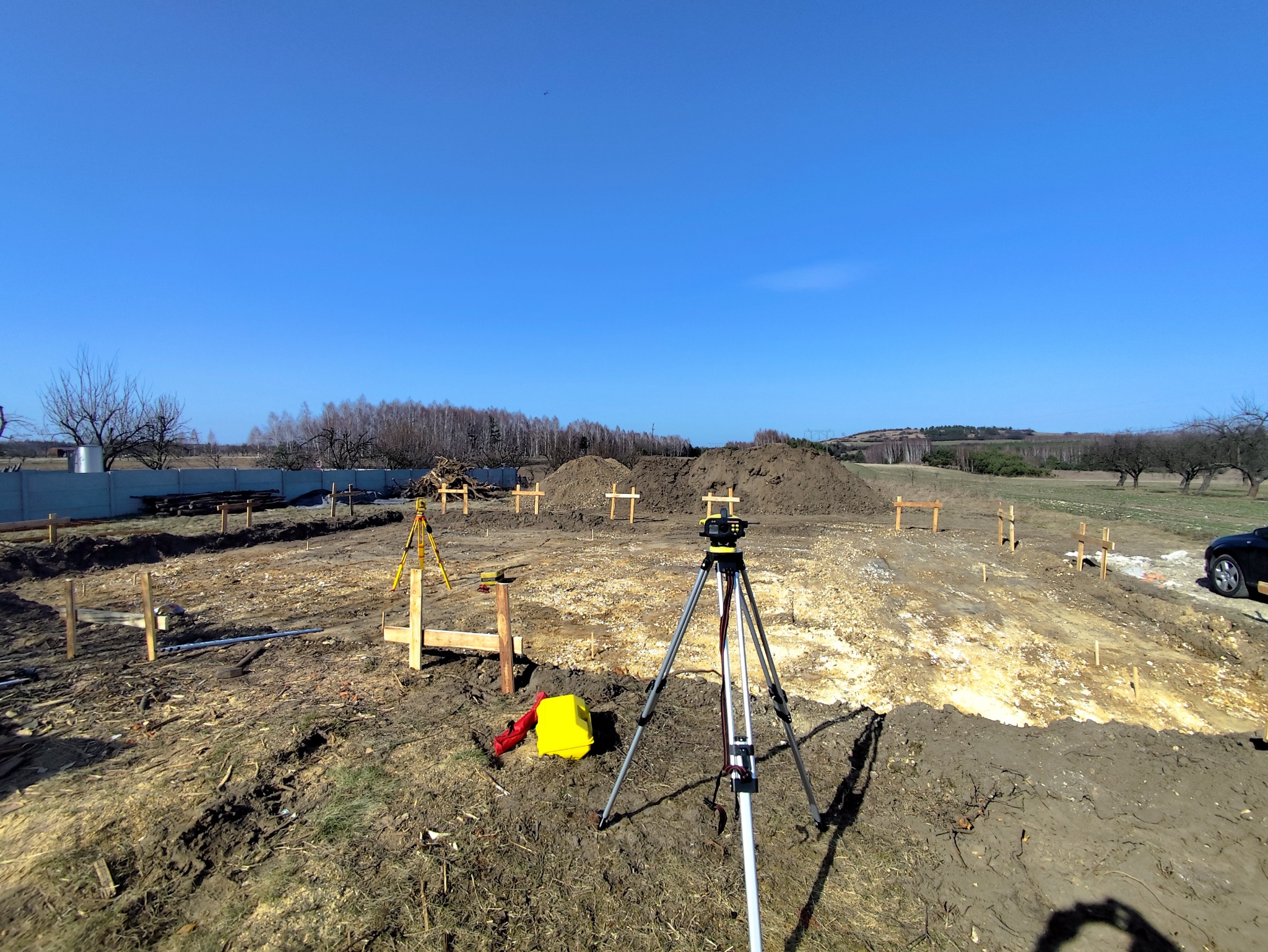 Geodezyjny pomiar terenu pod budowę z użyciem niwelatora i statywu. Wykop z drewnianymi palikami, żółta skrzynka narzędziowa, błękitne niebo.