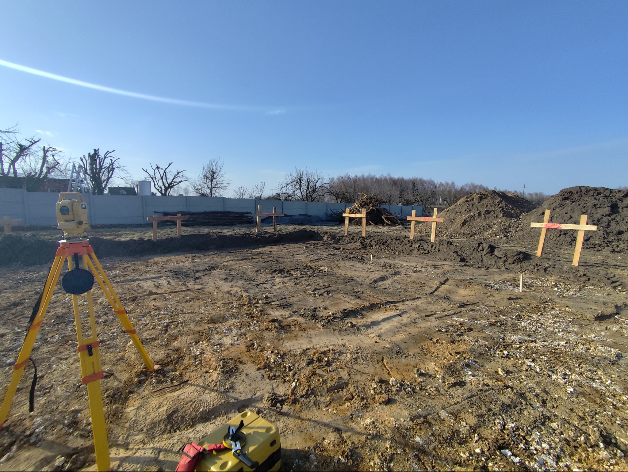 Żółty niwelator na statywie na placu budowy z wbitymi palikami. W tle widoczny szary płot i drzewa bez liści. Błękitne niebo.