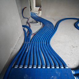 F.U.H. EFEKT ADAM GAWLAK, STANISŁAW GAWLAK SPÓŁKA CYWILNA - Niebieskie karbowane rury wentylacyjne ułożone równolegle na podłożu, przymocowane do niebieskiej płyty montażowej, instalacja w surowym stanie, widoczne ściany w trakcie wykończenia.