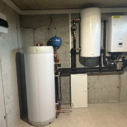 F.U.H. EFEKT ADAM GAWLAK, STANISŁAW GAWLAK SPÓŁKA CYWILNA - Zestaw urządzeń grzewczych w kotłowni: dwa białe zbiorniki, niebieskie naczynie przeponowe, piec gazowy marki Beretta oraz skrzynka elektryczna na szarej, surowej ścianie betonowej.