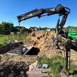 F.U.H. EFEKT ADAM GAWLAK, STANISŁAW GAWLAK SPÓŁKA CYWILNA - Montaż betonowego zbiornika pod ziemią za pomocą dźwigu samochodowego; pracownik wewnątrz studzienki kanalizacyjnej, wykop ziemny, widoczny krajobraz wiejski w tle.