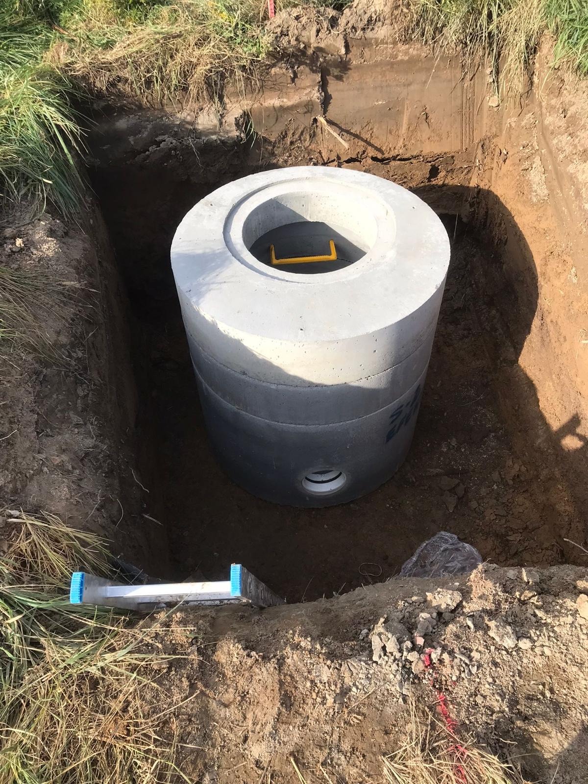 Betonowy zbiornik na szambo lub deszczówkę umieszczony w wykopie, widoczny poziomica na krawędzi wykopu i czerwona taśma geodezyjna.