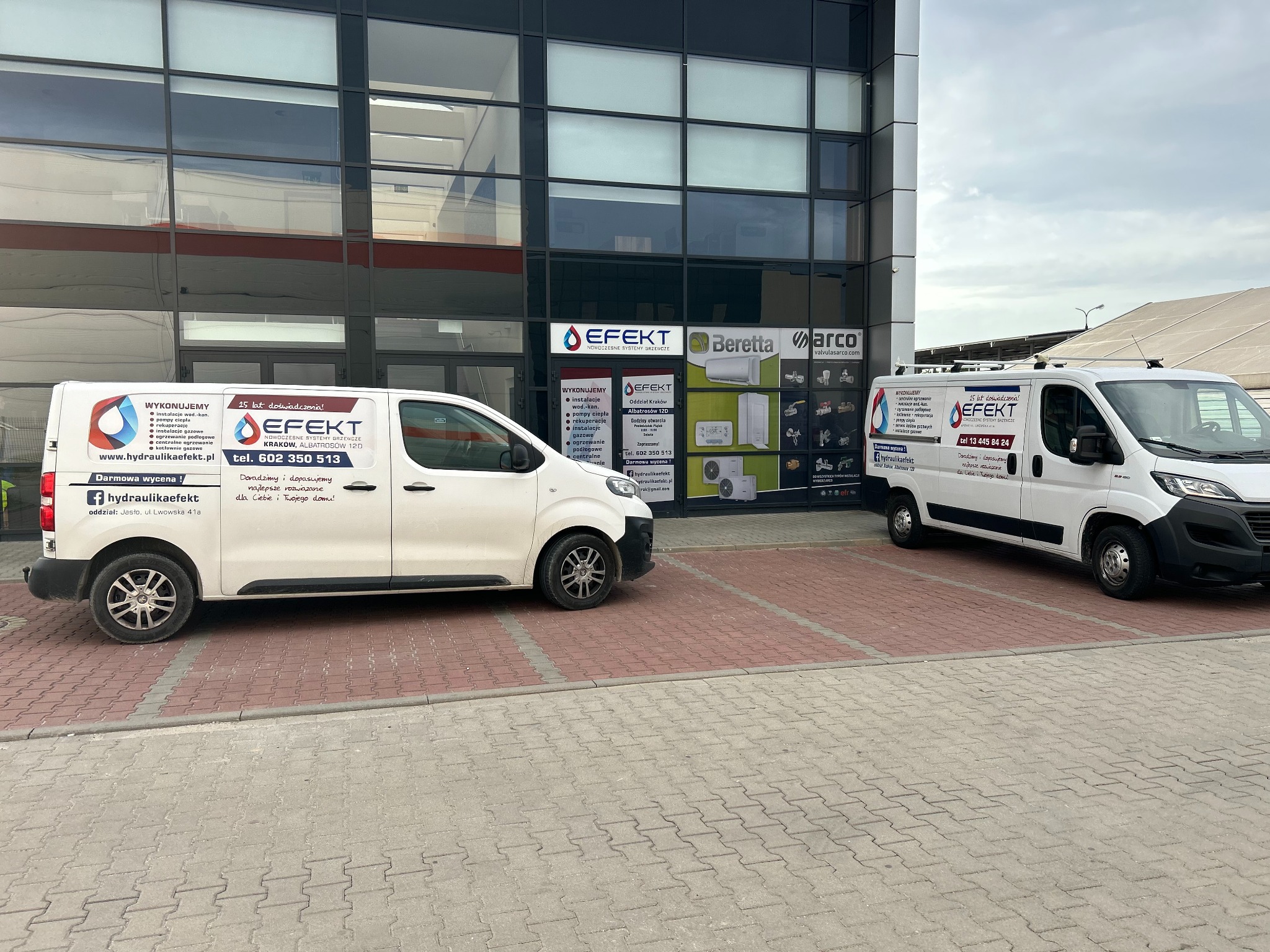 Dwa białe samochody dostawcze firmy instalacyjnej EFEKT zaparkowane przed nowoczesnym budynkiem biurowym z dużymi oknami, na których widoczne są logotypy producentów urządzeń grzewczych.