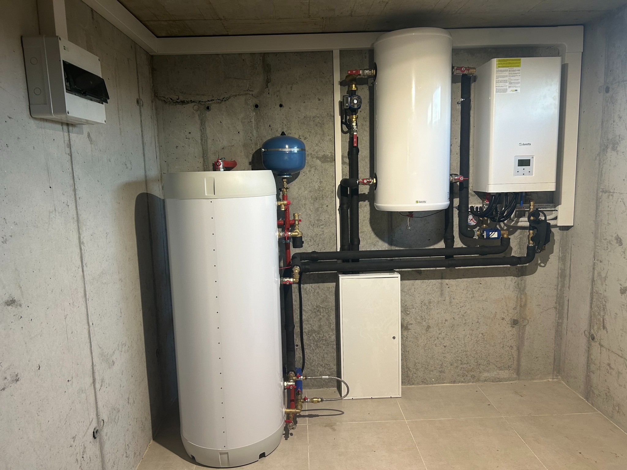 Zestaw urządzeń grzewczych w kotłowni: dwa białe zbiorniki, niebieskie naczynie przeponowe, piec gazowy marki Beretta oraz skrzynka elektryczna na szarej, surowej ścianie betonowej.