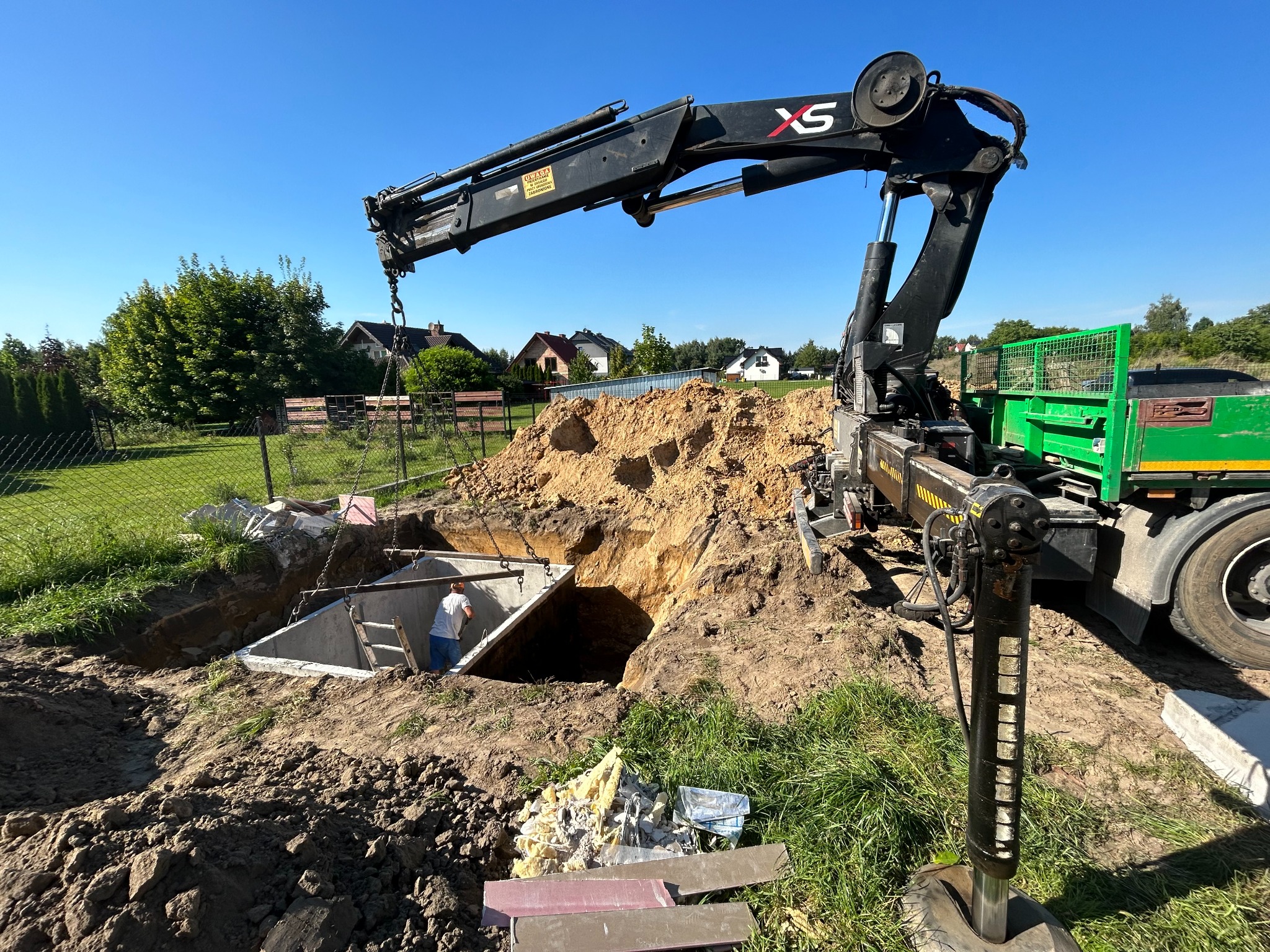 Montaż betonowego zbiornika pod ziemią za pomocą dźwigu samochodowego; pracownik wewnątrz studzienki kanalizacyjnej, wykop ziemny, widoczny krajobraz wiejski w tle.