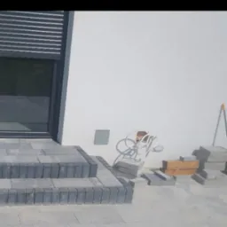 Układanie kostki brukowej na schodach wejściowych do domu z szarą elewacją, widoczne kable wystające ze ściany.