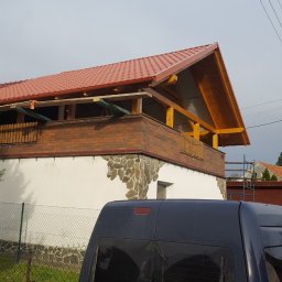 Roman Gadziała - Dom z nowym, czerwonym dachem z blachodachówki i drewnianą konstrukcją balkonu, widoczny zza zaparkowanego, brudnego samochodu dostawczego z uszkodzoną szybą, w pochmurny dzień.