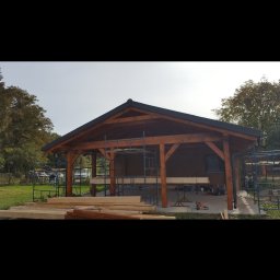 Roman Gadziała - Drewniana konstrukcja z dachem w trakcie budowy, widoczne rusztowania, stos drewna i narzędzia na zewnątrz budynku.