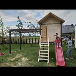 Roman Gadziała - Drewniany domek dla dzieci ze schodami i czerwoną zjeżdżalnią, na trawniku, w tle pergola i zadbany ogród, dzieci i dorosły w pobliżu.