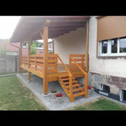 Drewniana altana z zadaszeniem, schodami i balustradą, otoczona kamieniami i kwiatami w donicach, przylegająca do budynku z roletami zewnętrznymi.