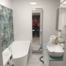 Roman Gadziała - Wnętrze łazienki po remoncie: biała wanna narożna, pralka, umywalka nablatowa na szafce z szufladami, lustro w wąskiej ramie, płytki dekoracyjne na ścianie przy wannie oraz szare płytki na podłodze.