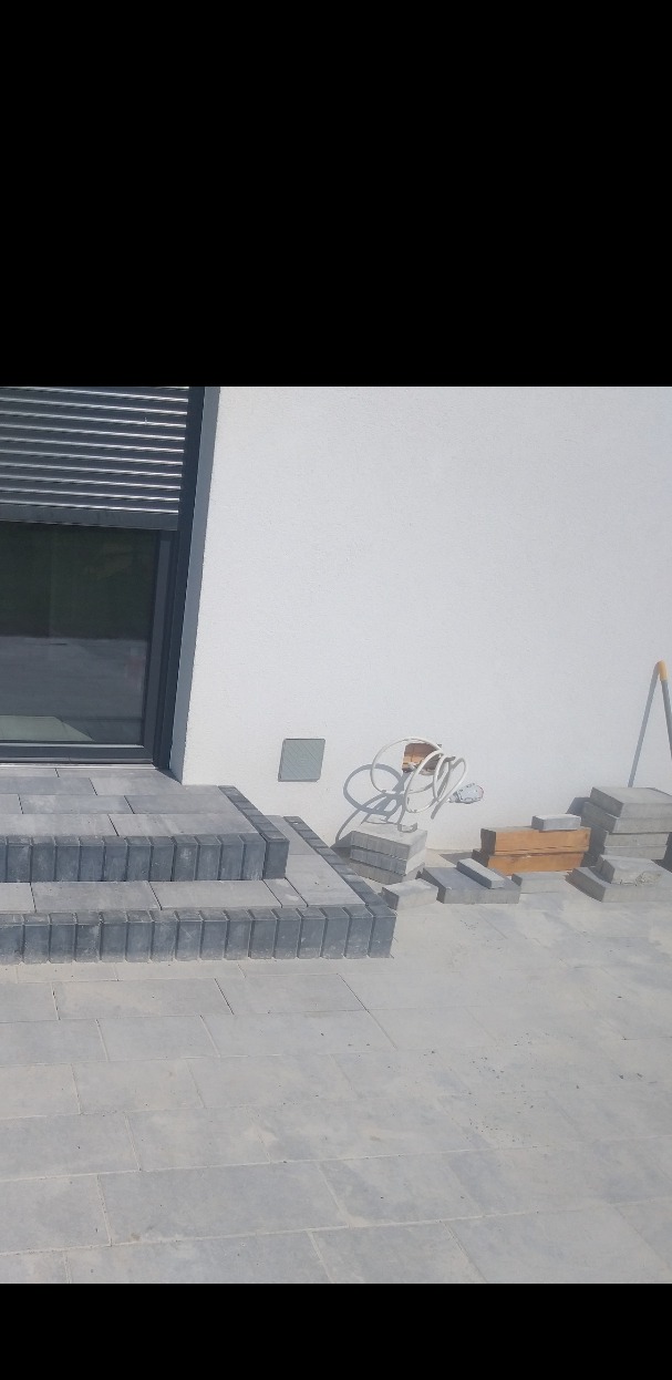 Układanie kostki brukowej na schodach wejściowych do domu z szarą elewacją, widoczne kable wystające ze ściany.