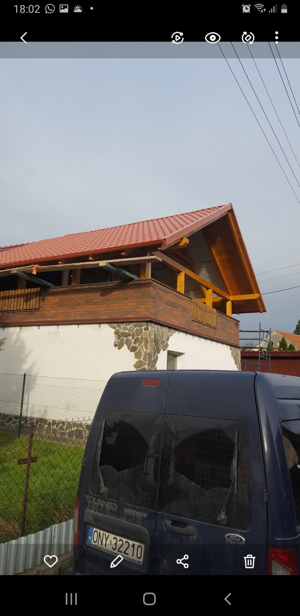 Dom z nowym, czerwonym dachem z blachodachówki i drewnianą konstrukcją balkonu, widoczny zza zaparkowanego, brudnego samochodu dostawczego z uszkodzoną szybą, w pochmurny dzień.