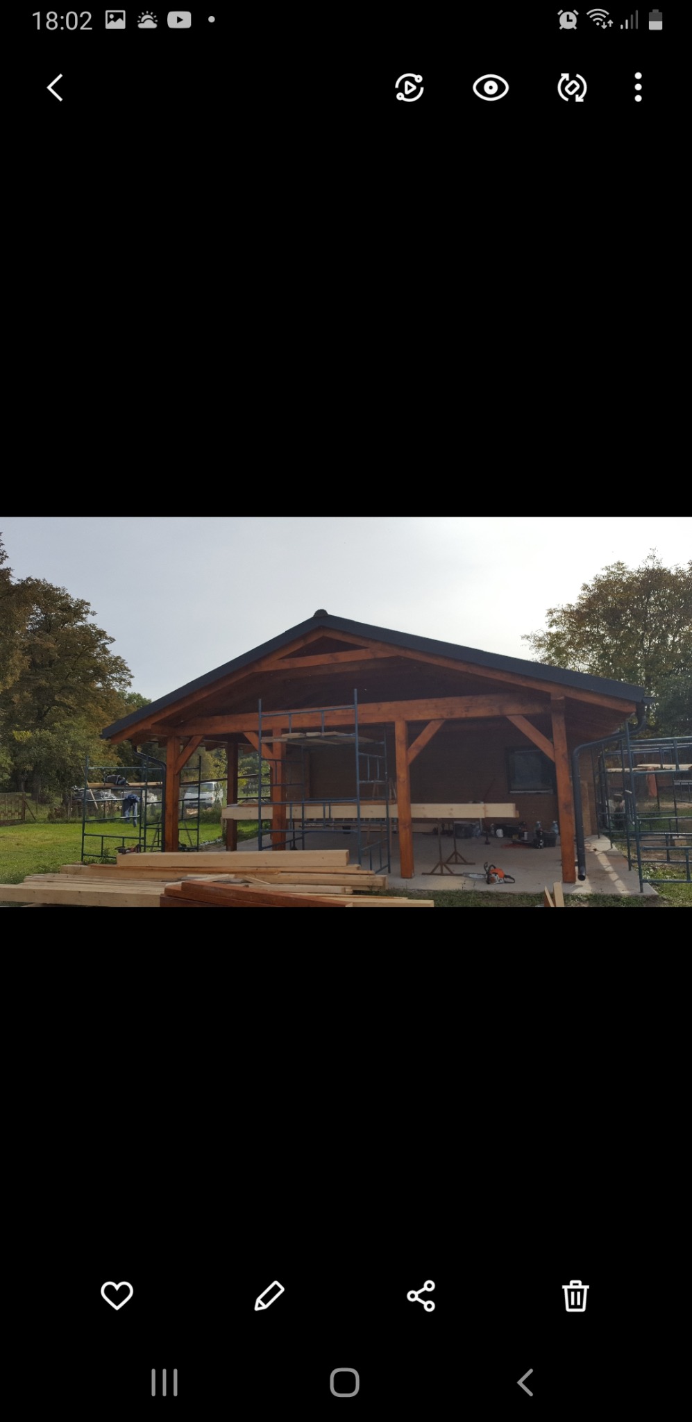 Drewniana konstrukcja z dachem w trakcie budowy, widoczne rusztowania, stos drewna i narzędzia na zewnątrz budynku.