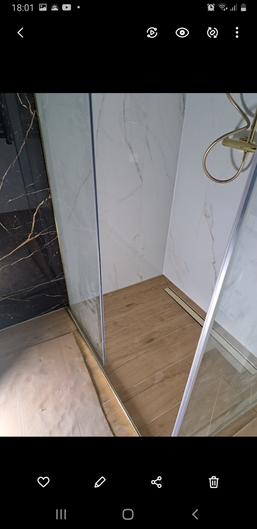 Nowoczesna kabina prysznicowa z transparentnymi drzwiami, złotymi akcentami i odpływem liniowym, wykończona płytkami imitującymi drewno i marmur.