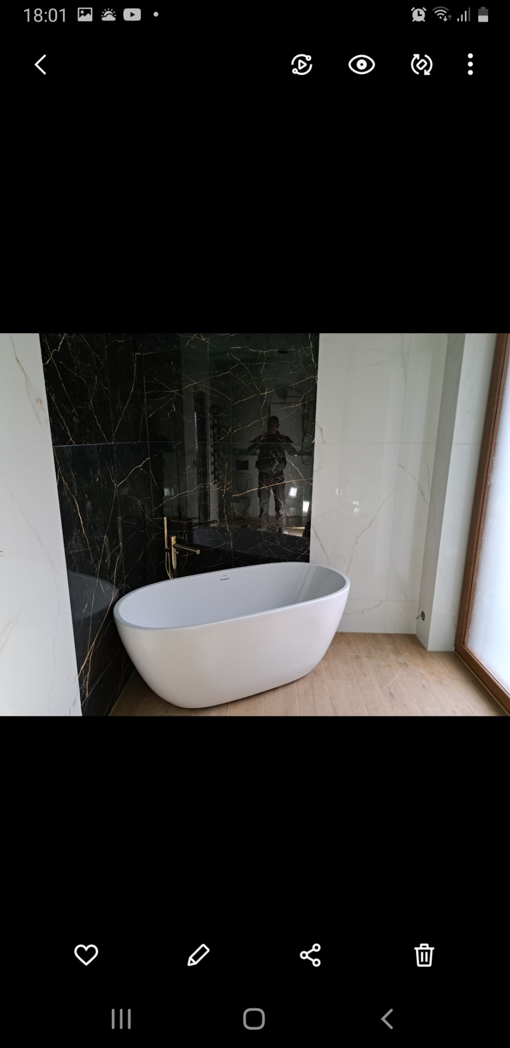 Nowoczesna, wolnostojąca wanna owalna z złotą armaturą na tle czarno-białych marmurowych płytek w wykończonej łazience, widoczne odbicie osoby w lustrzanej powierzchni płytek.