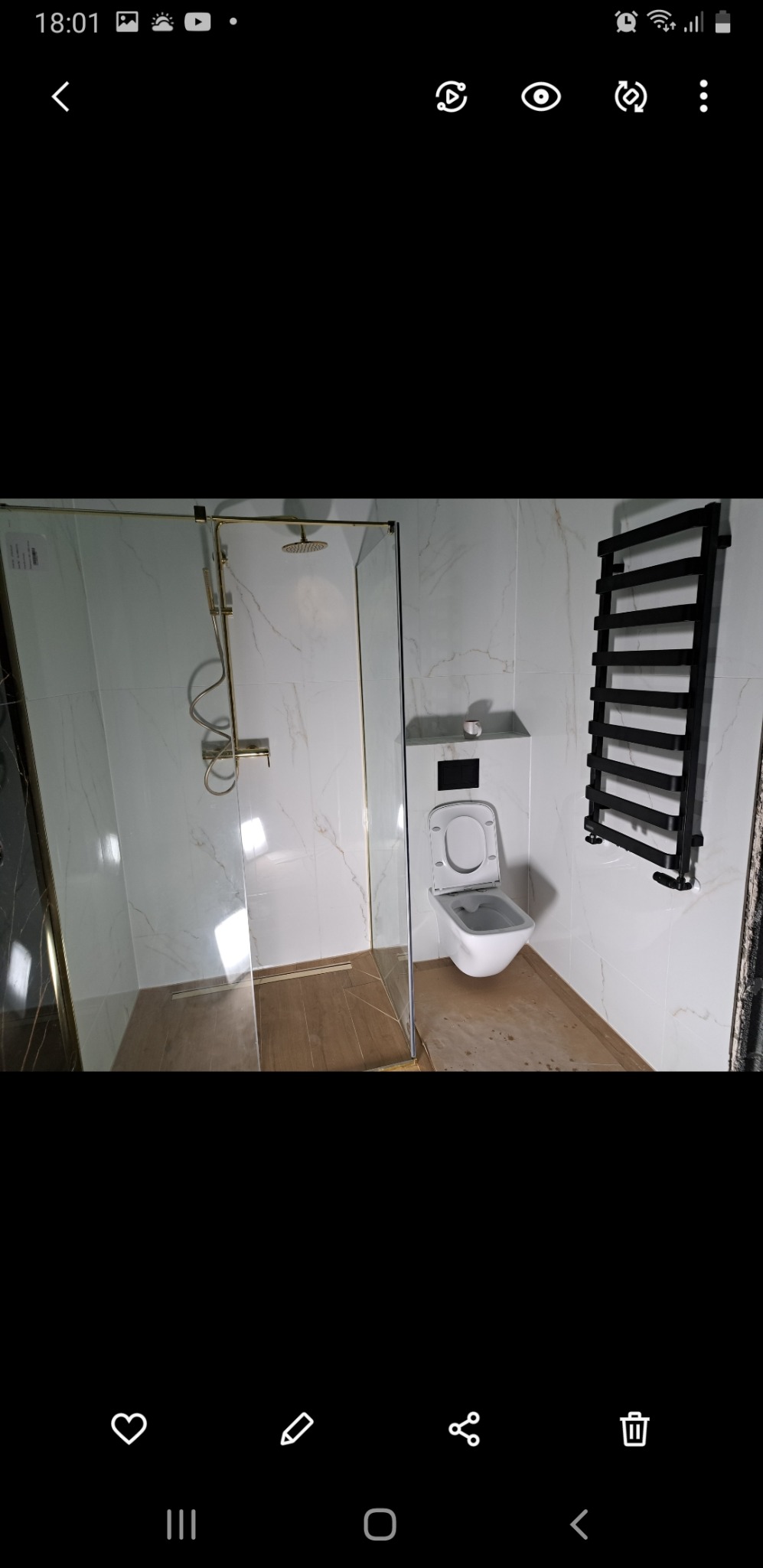 Nowoczesna łazienka z kabiną prysznicową ze złotymi elementami, podwieszaną toaletą i czarnym grzejnikiem drabinkowym na marmurowych płytkach.