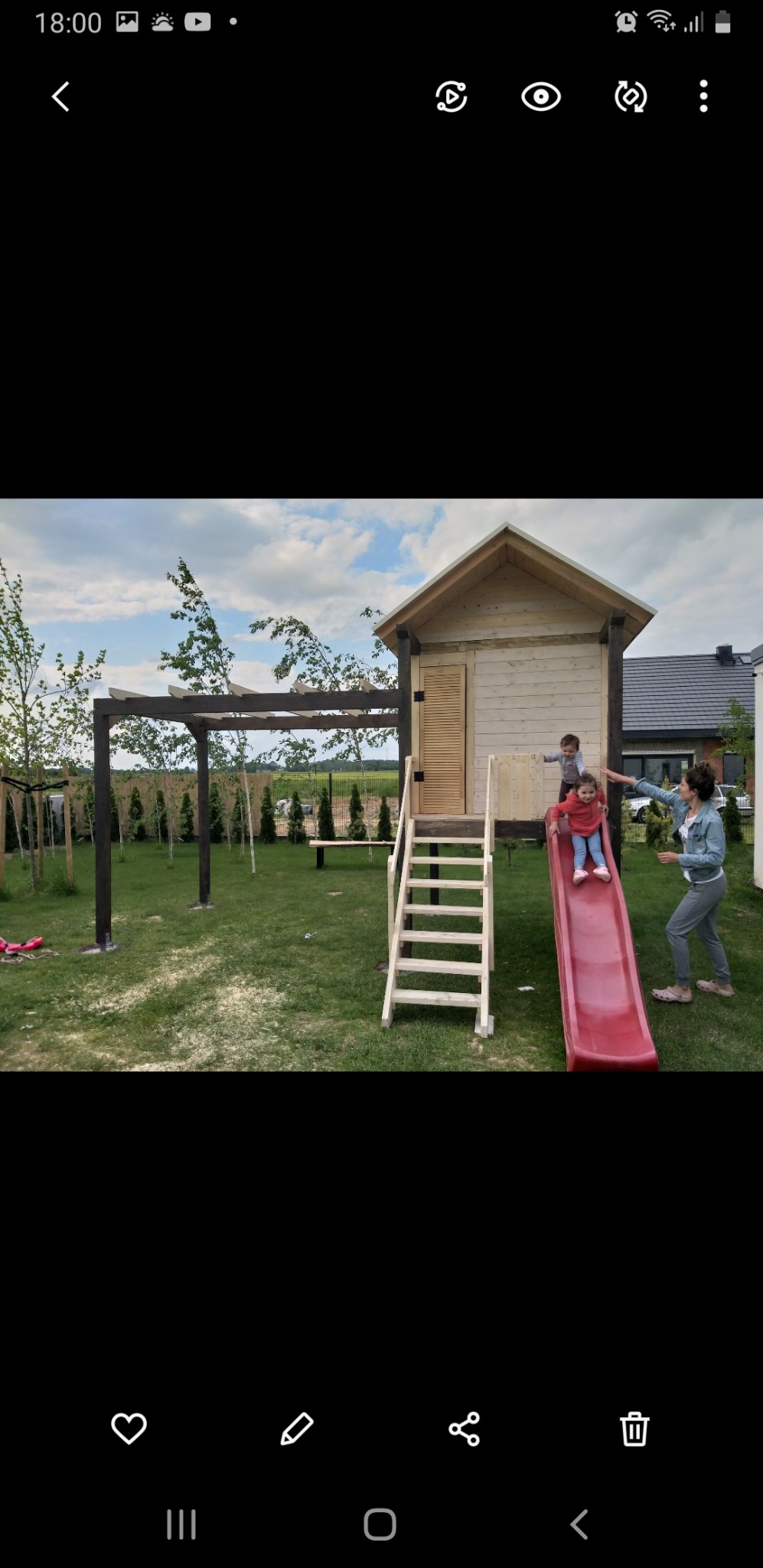 Drewniany domek dla dzieci ze schodami i czerwoną zjeżdżalnią, na trawniku, w tle pergola i zadbany ogród, dzieci i dorosły w pobliżu.