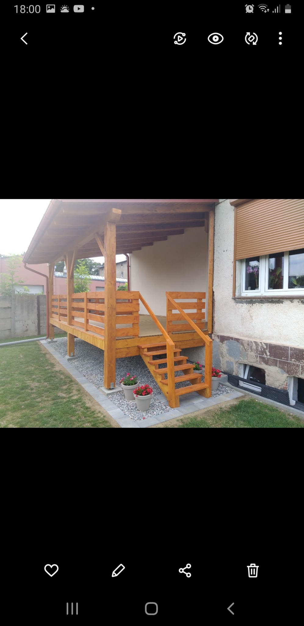 Drewniana altana z zadaszeniem, schodami i balustradą, otoczona kamieniami i kwiatami w donicach, przylegająca do budynku z roletami zewnętrznymi.