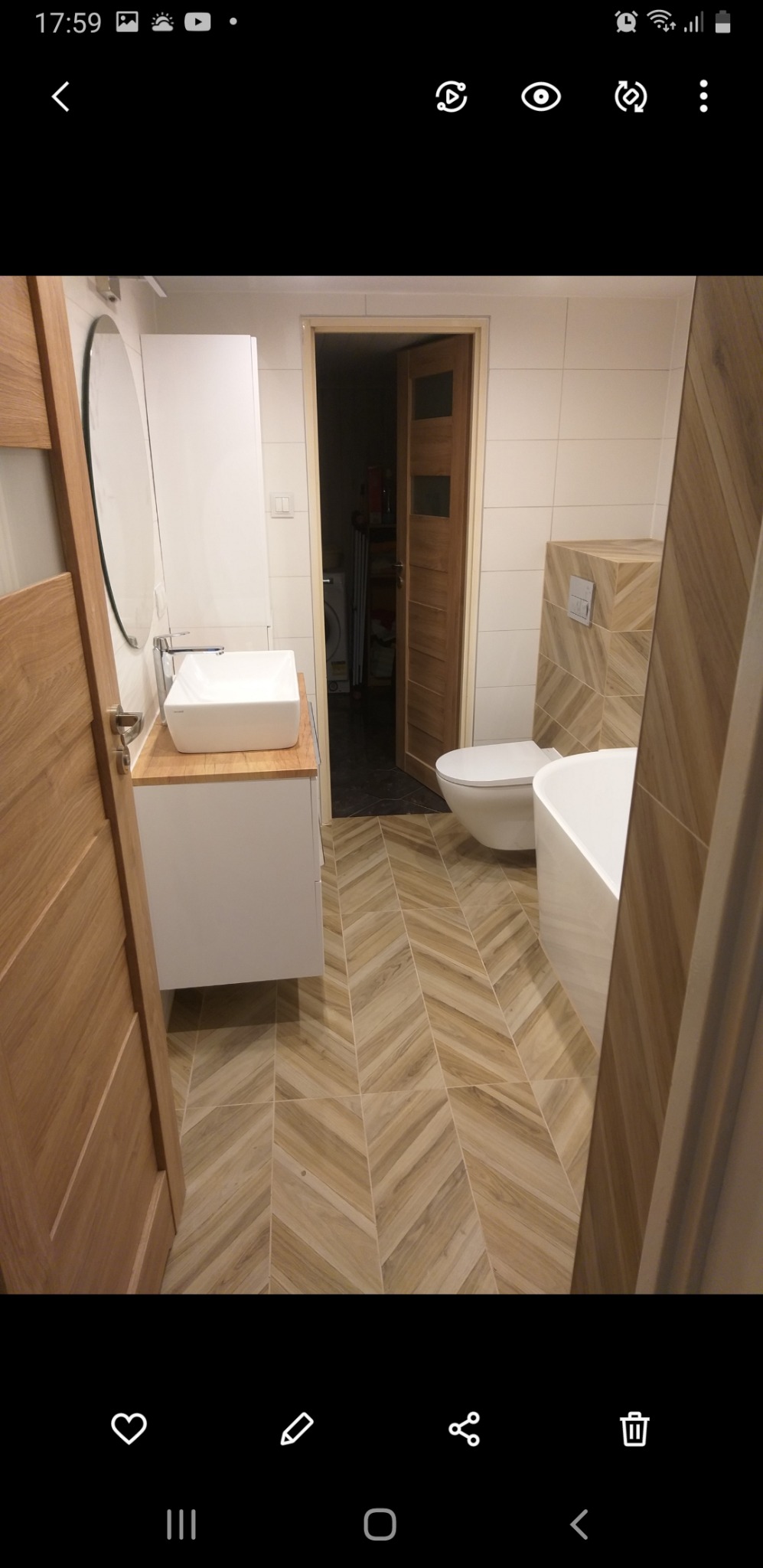 Nowoczesna łazienka z białą armaturą, drewnopodobną glazurą ułożoną w jodełkę na podłodze i ścianie, oraz drewnianymi drzwiami.