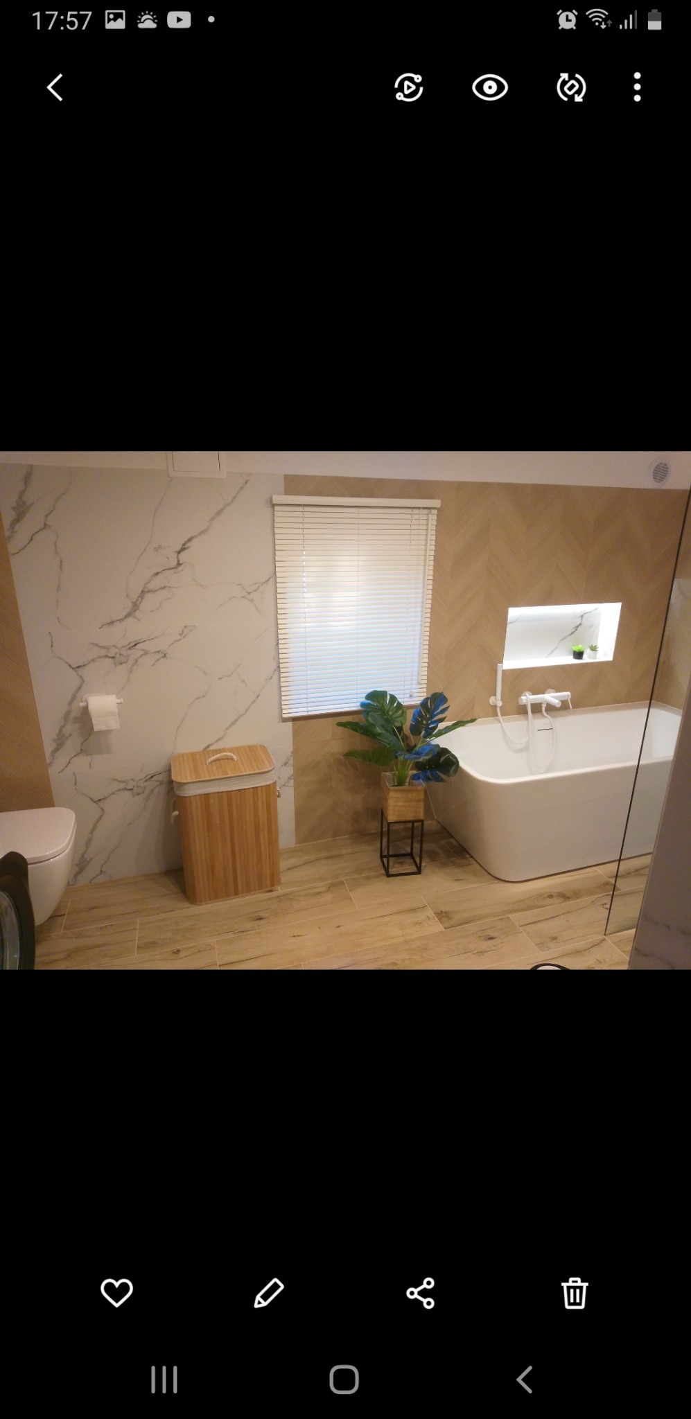 Nowoczesna łazienka z białymi płytkami imitującymi marmur na ścianie, drewnianą podłogą, białą wanną, drewnianym koszem na pranie i roletami w oknie, dopełniona rośliną doniczkową.