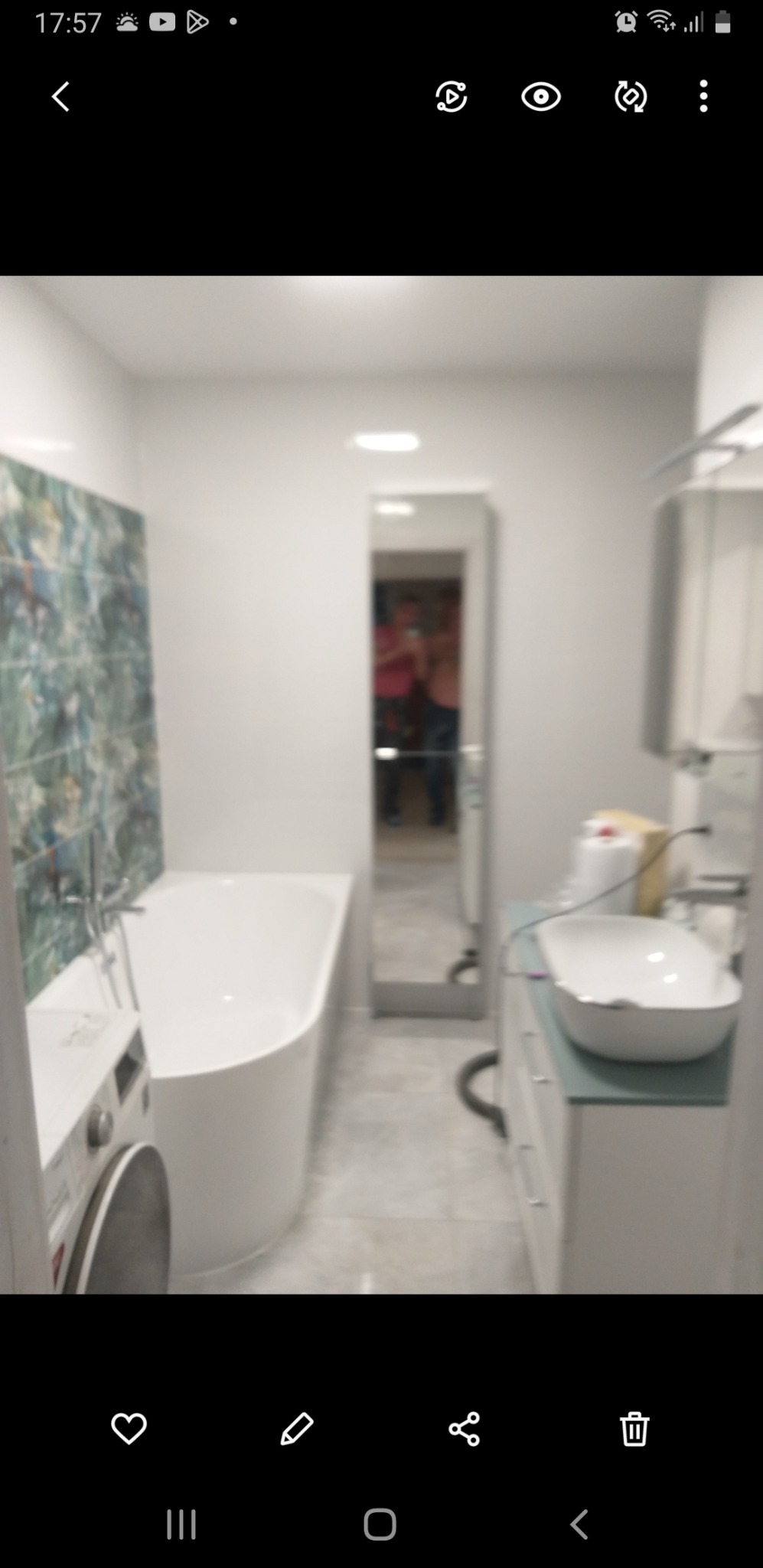 Wnętrze łazienki po remoncie: biała wanna narożna, pralka, umywalka nablatowa na szafce z szufladami, lustro w wąskiej ramie, płytki dekoracyjne na ścianie przy wannie oraz szare płytki na podłodze.