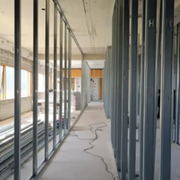 Metalowa konstrukcja szkieletowa ścian działowych w surowym wnętrzu, widoczne profile pionowe z wycięciami w kształcie litery H, przygotowane do obłożenia płytami, z oknami w tle po lewej stronie...