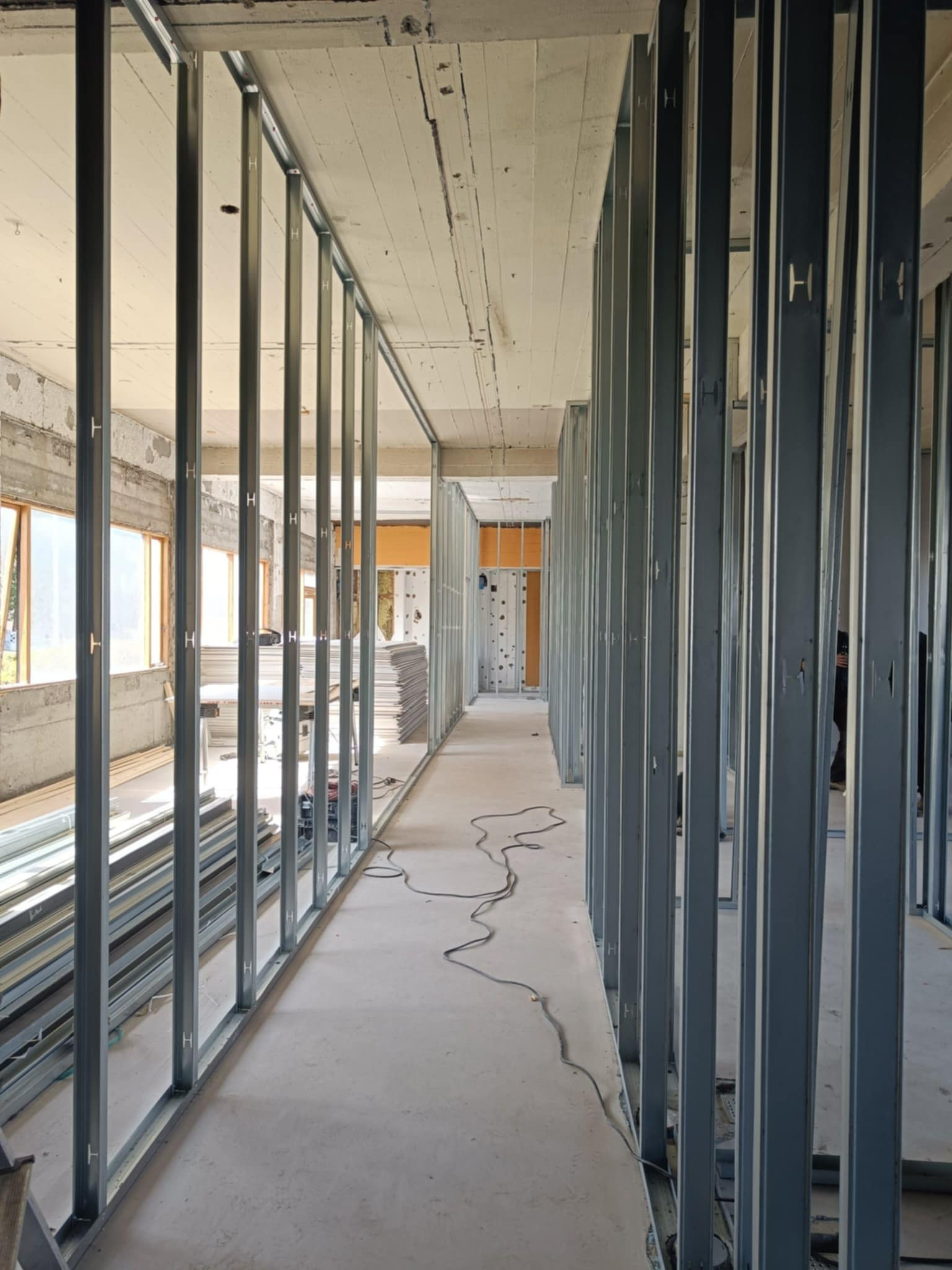 Metalowa konstrukcja szkieletowa ścian działowych w surowym wnętrzu, widoczne profile pionowe z wycięciami w kształcie litery H, przygotowane do obłożenia płytami, z oknami w tle po lewej stronie...