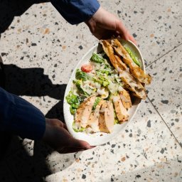 Trifolati Społka z O.O. - Sałatka Cezar z kurczakiem i grzankami, trzymana przez osobę w niebieskiej koszuli, na tle lastryko w plamach światła i cienia.