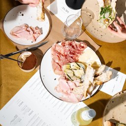 Trifolati Społka z O.O. - Stół z włoskimi przekąskami: wędliny, sery, pizza, tiramisu, menu restauracji Andiamo, napoje. Widok z góry, ciepłe oświetlenie, ręce sięgające po jedzenie.