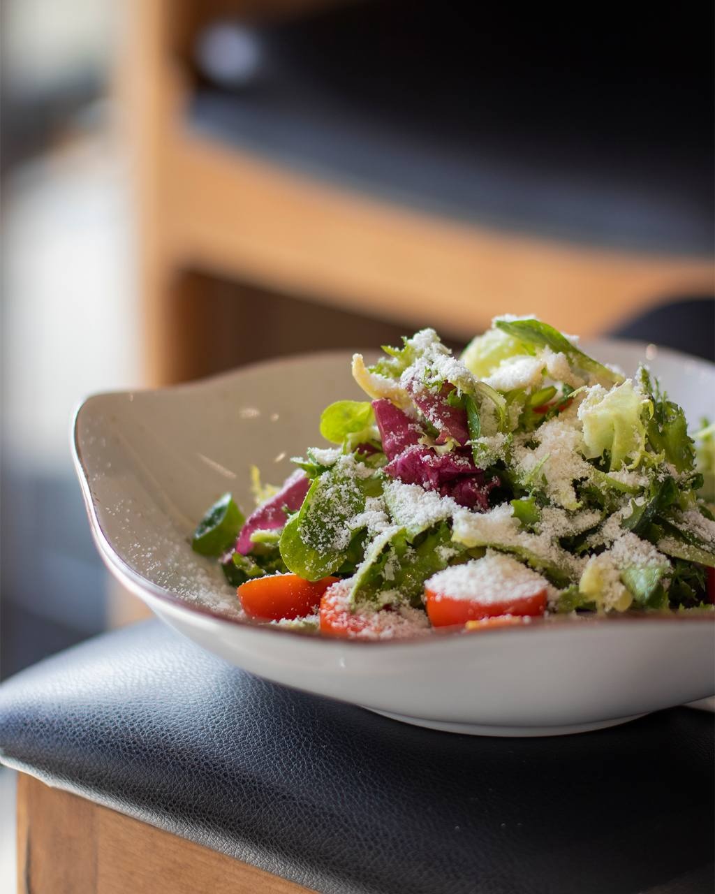 Świeża sałatka z pomidorkami koktajlowymi i posypką serową w białej, designerskiej misce, ustawiona na czarnym skórzanym siedzisku krzesła.