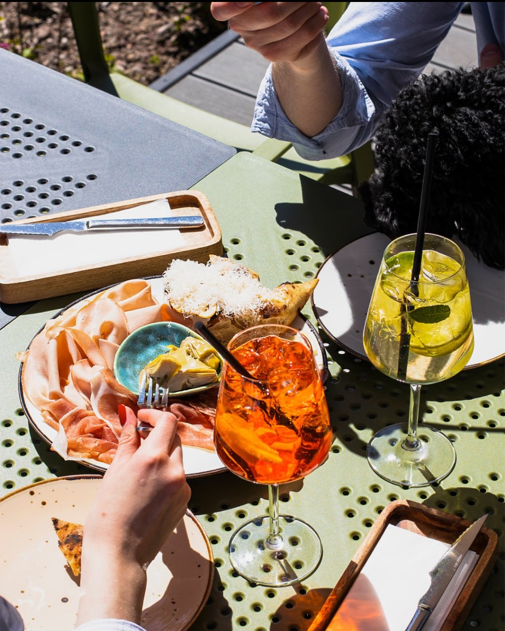 Aranżacja stołu z przekąskami: szynka, pieczywo z serem, karczochy i dwa drinki na zielonym stole z metalowymi elementami w słoneczny dzień.