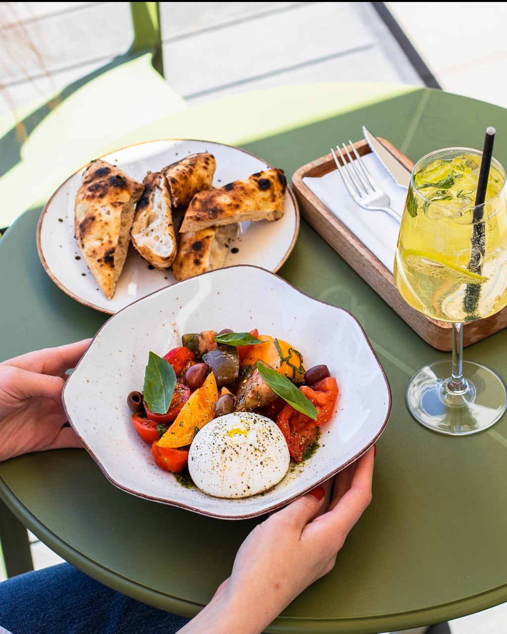 Sałatka z burratą i warzywami w dłoniach, obok pieczywo i drink na zielonym stole. Jasne, naturalne światło.