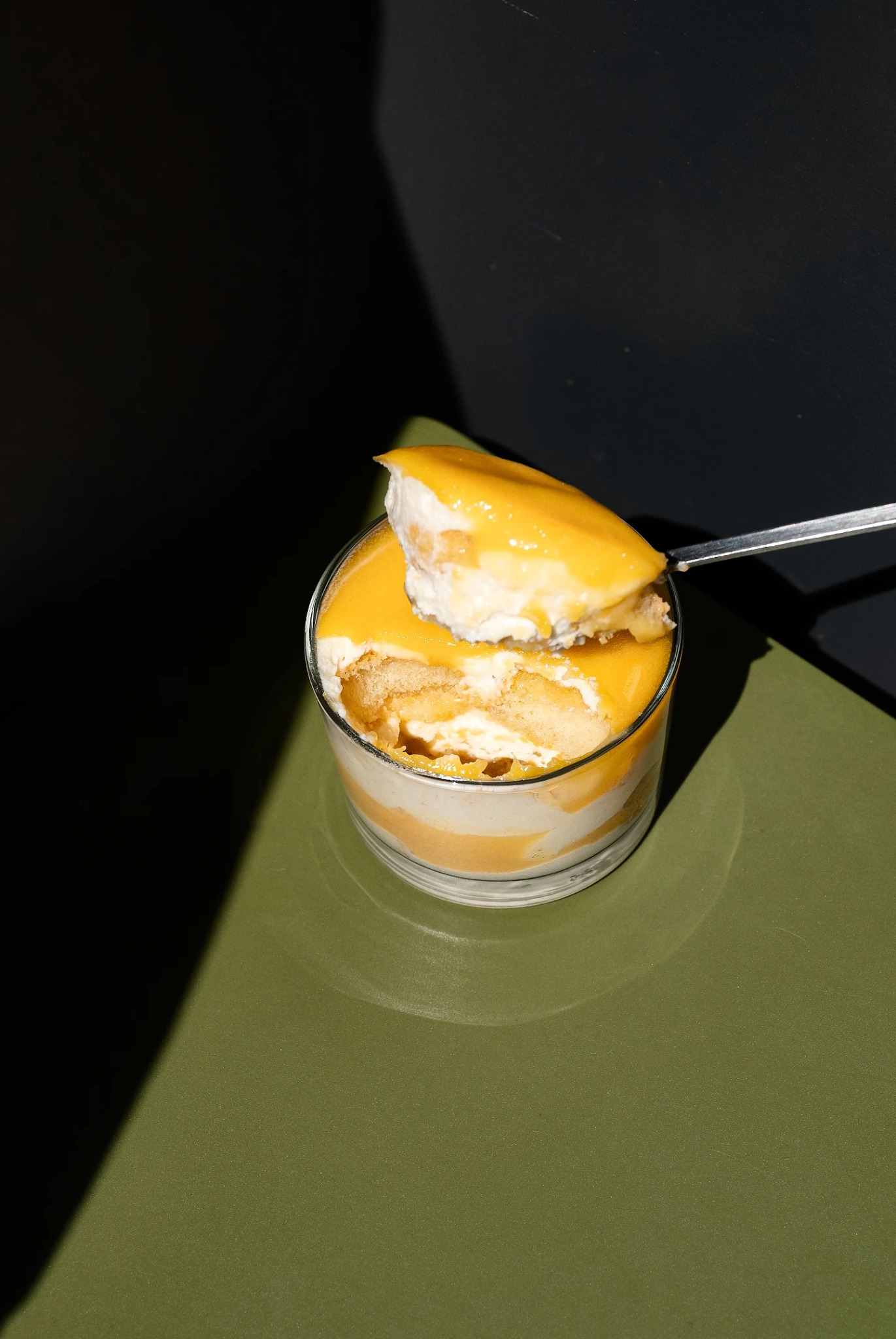 Deser w szklance, warstwy kremu i mango, łyżeczka nabiera porcję. Minimalistyczne ujęcie z cieniem na zielonym tle, nowoczesny styl.