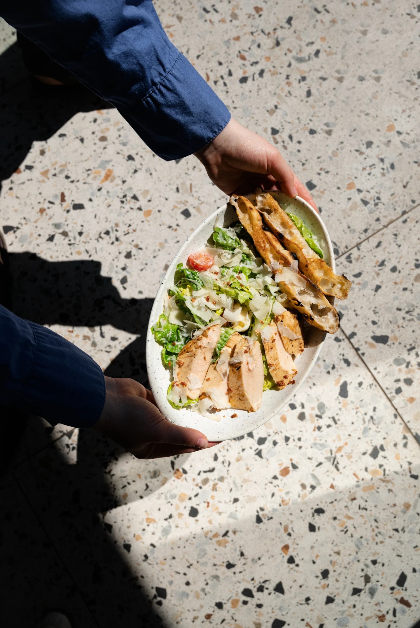 Sałatka Cezar z kurczakiem i grzankami, trzymana przez osobę w niebieskiej koszuli, na tle lastryko w plamach światła i cienia.