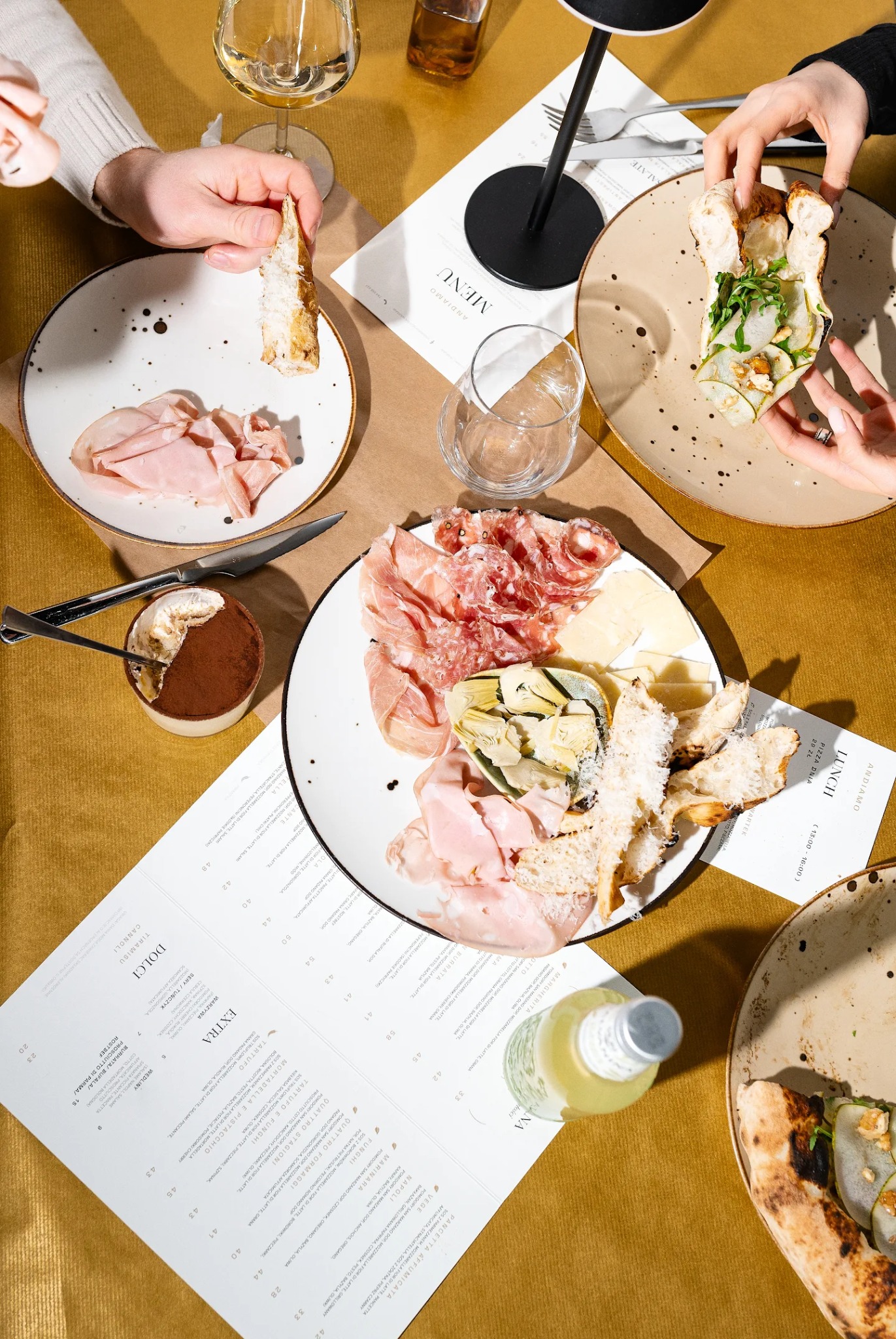 Stół z włoskimi przekąskami: wędliny, sery, pizza, tiramisu, menu restauracji Andiamo, napoje. Widok z góry, ciepłe oświetlenie, ręce sięgające po jedzenie.