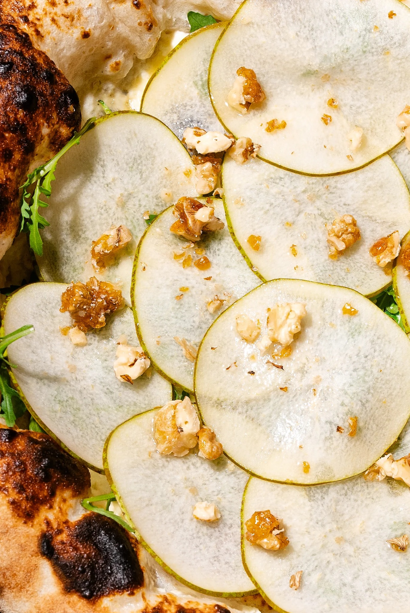 Pizza z gruszką i orzechami włoskimi, z bliska. Delikatne plastry gruszki ułożone na cieście, posypane orzechami w karmelu. Widoczne listki rukoli.