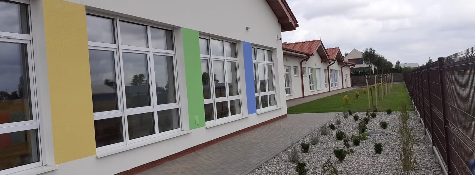 Budynek przedszkola z kolorowymi akcentami na elewacji, widoczne okna, chodnik z kostki brukowej, trawnik i ogrodzenie z siatki.