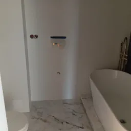 Wnętrze łazienki w trakcie prac wykończeniowych:  wolnostojąca wanna, marmurowe płytki podłogowe, ściany przygotowane pod montaż armatury, widoczne puszki elektryczne i  taśma malarska...