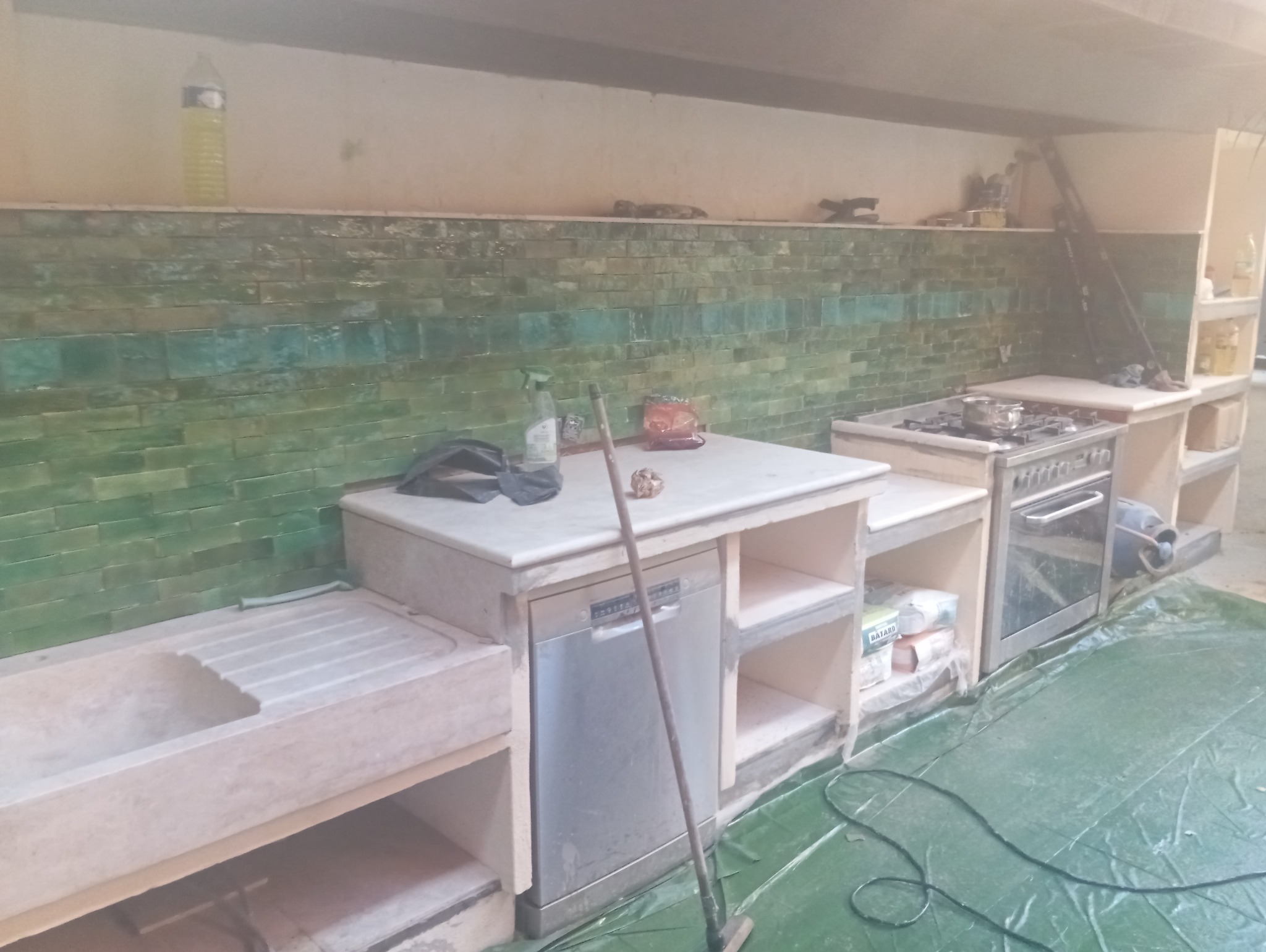 Wnętrze kuchni w trakcie remontu, widoczna zielona ściana z płytek, zabudowa kuchenna z kamienia, kuchenka gazowa i zmywarka, na podłodze folia ochronna.