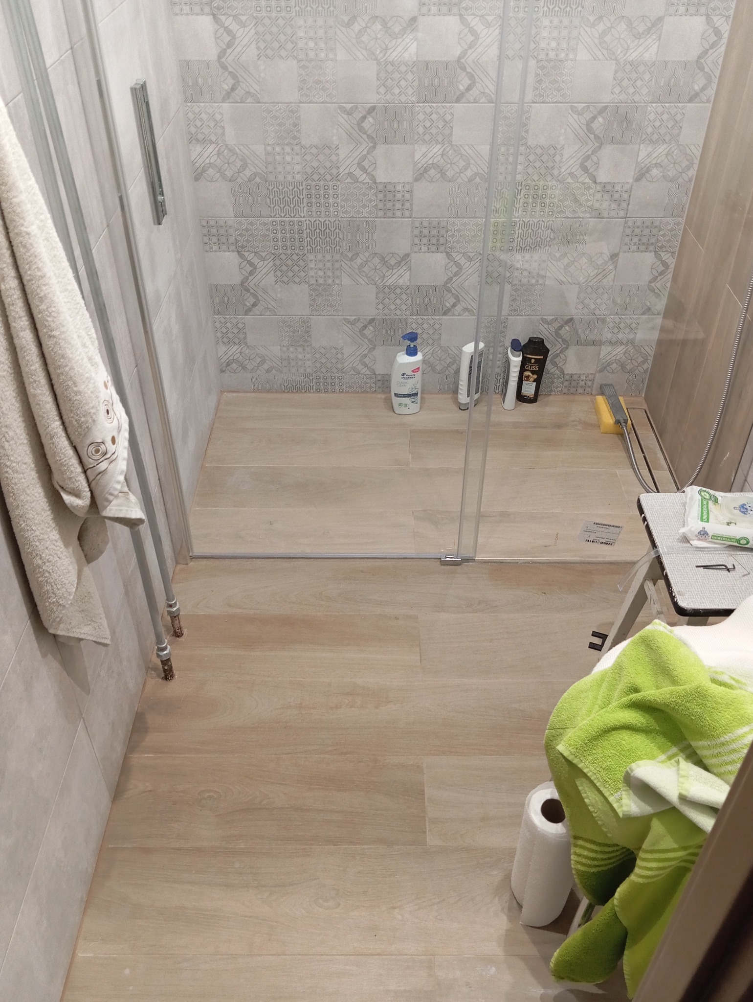 Nowoczesna kabina prysznicowa z jasnymi płytkami imitującymi drewno na podłodze i wzorzystymi szarymi płytkami na ścianach, widoczne butelki z kosmetykami pod prysznicem, ręcznik na wieszaku...