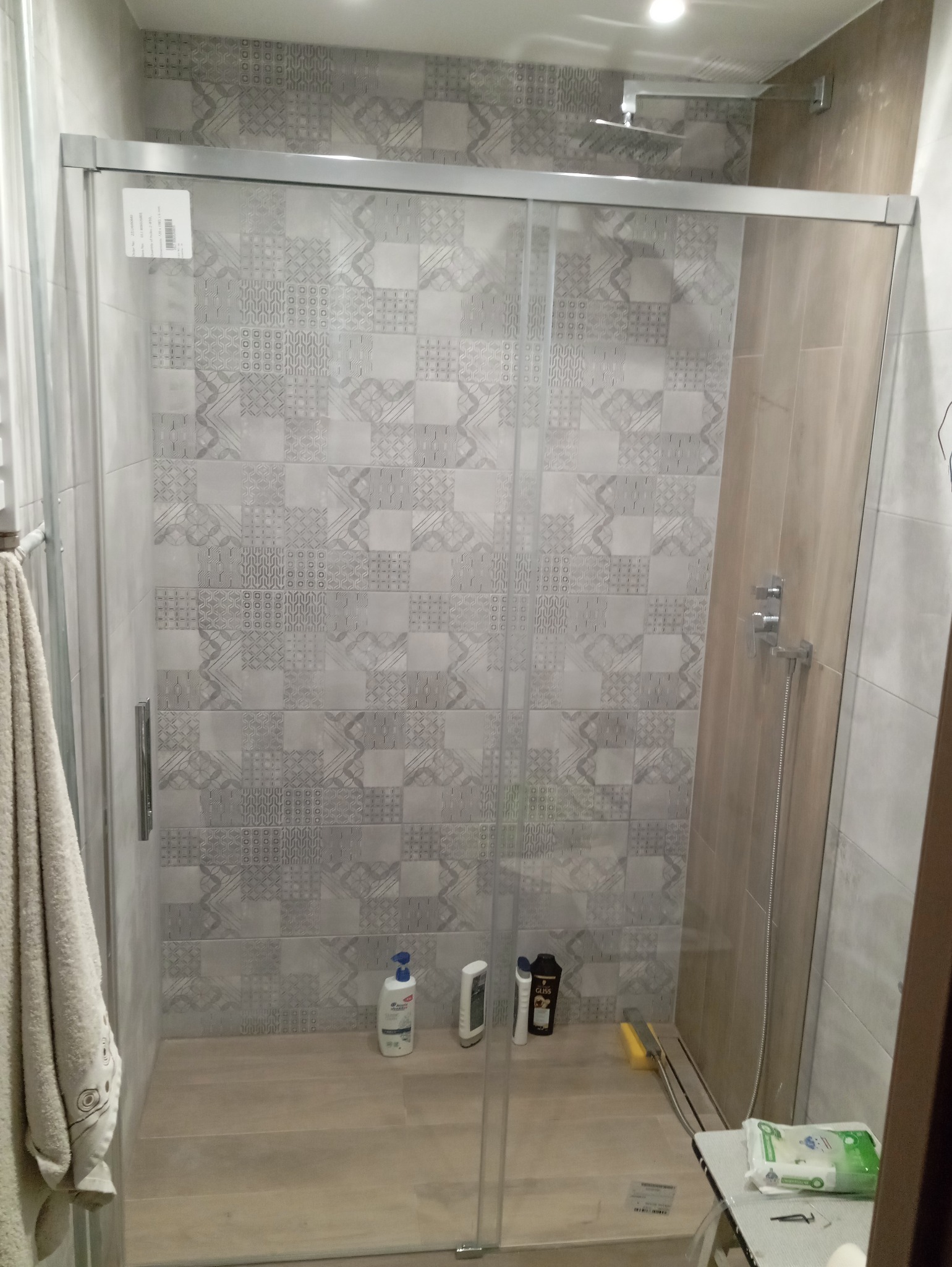 Szklana kabina prysznicowa z przesuwnymi drzwiami, szare płytki z wzorami na ścianach, ręcznik na relingu po lewej, kosmetyki pod prysznicem, odpływ liniowy przy ścianie.