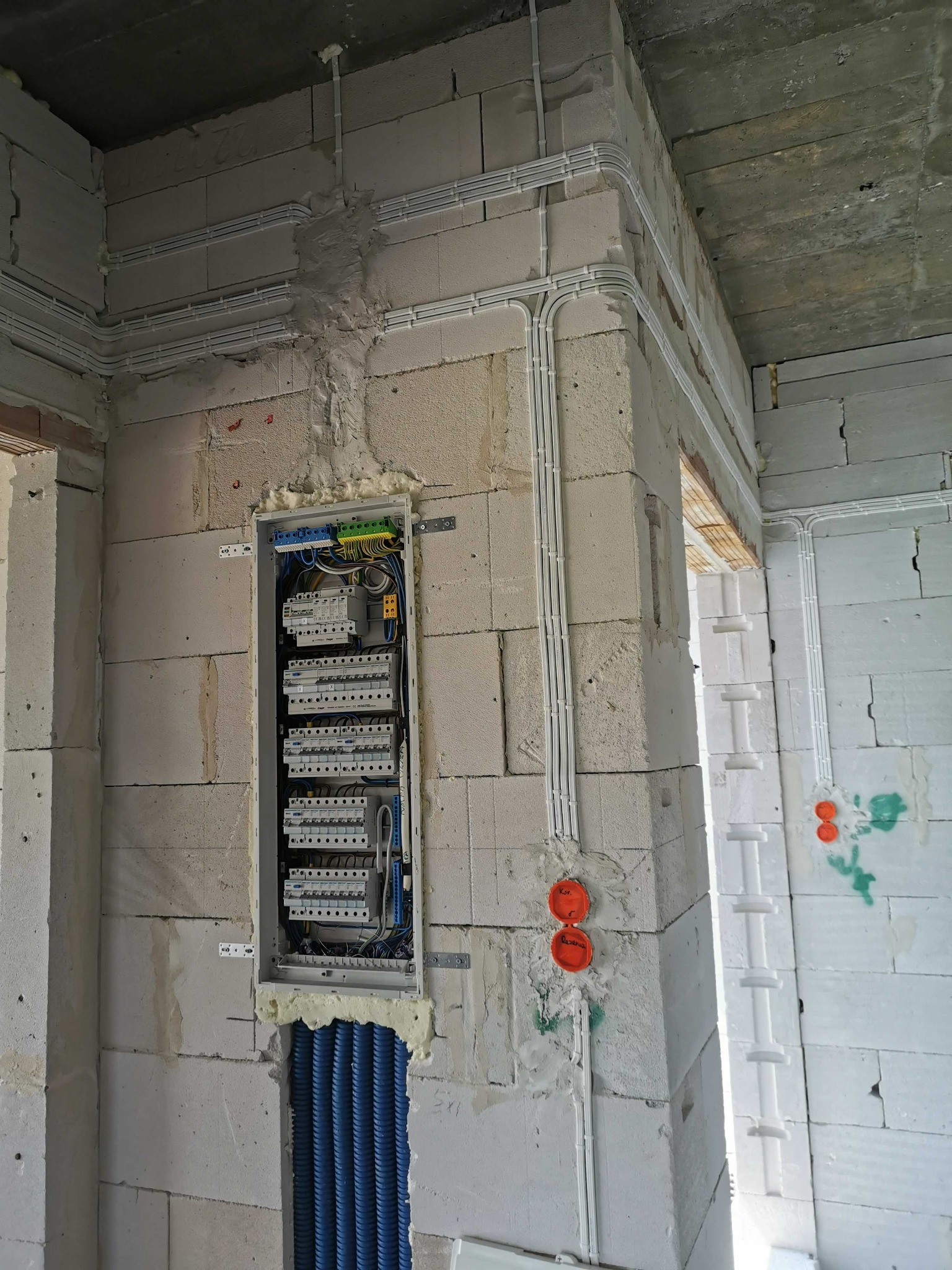 Rozdzielnia elektryczna w surowym stanie, widoczne białe kable w peszlach na ścianie z bloczków, niebieskie rury karbowane pod rozdzielnicą, ślady pianki montażowej.