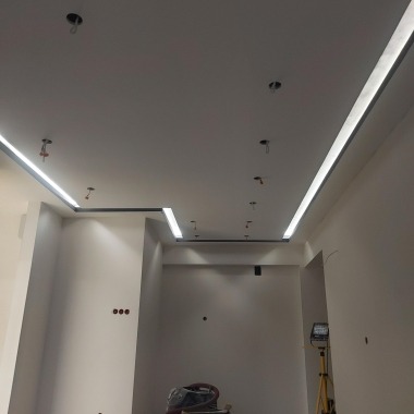 Biały sufit podwieszany z wbudowanym oświetleniem LED w kształcie litery L, widoczne otwory na dodatkowe punkty świetlne i wystające kable, lampa robocza na statywie w rogu pomieszczenia.