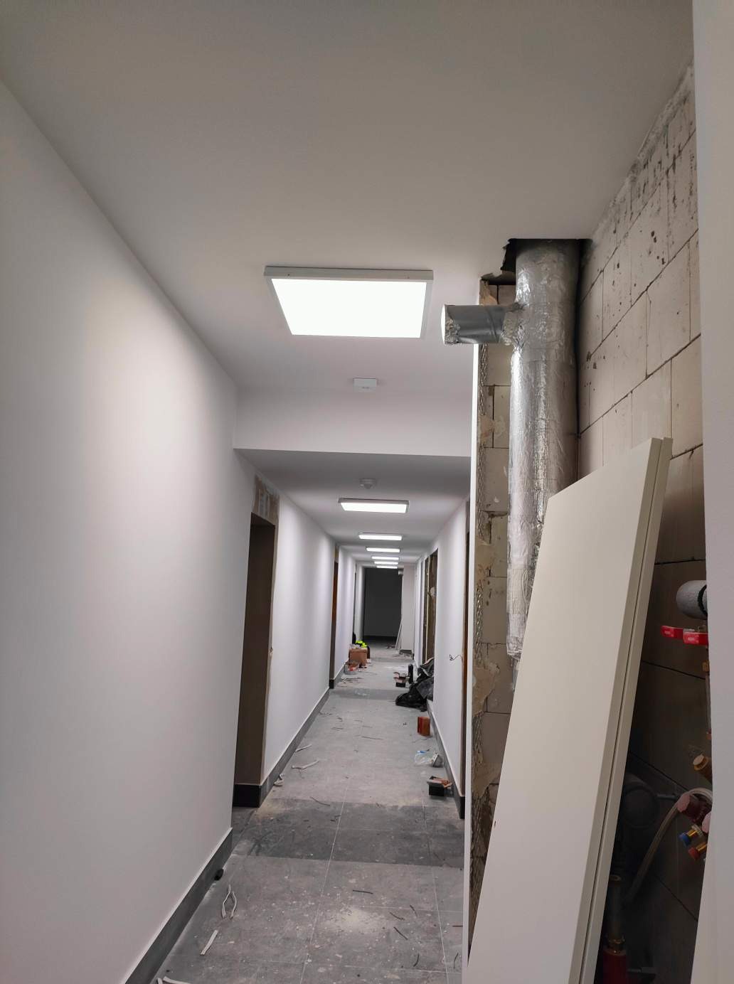 Korytarz w trakcie remontu z zamontowanym oświetleniem LED, widoczny przewód wentylacyjny w izolacji aluminiowej i elementy instalacji hydraulicznej.