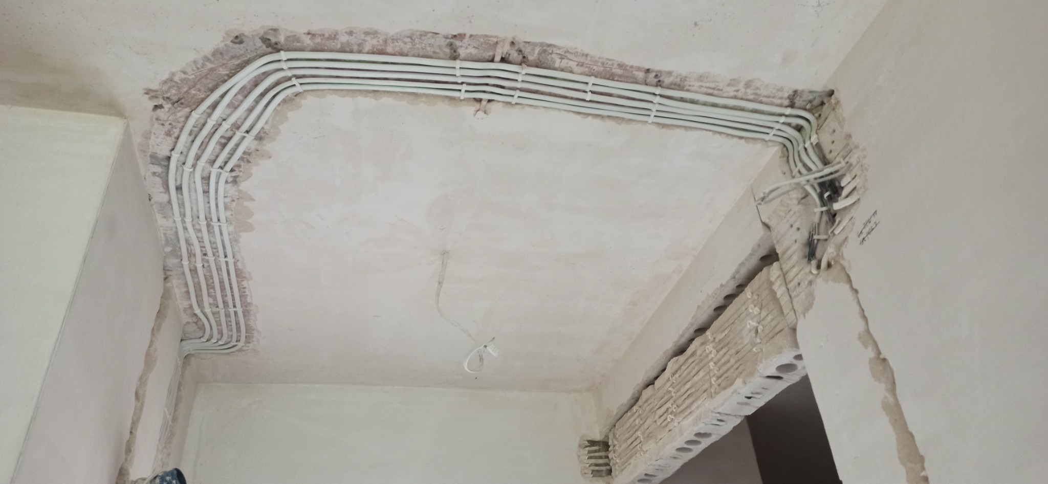 Białe rurki instalacji elektrycznej ułożone wzdłuż sufitu w bruzdach, zabezpieczone uchwytami, sufit w trakcie prac wykończeniowych.