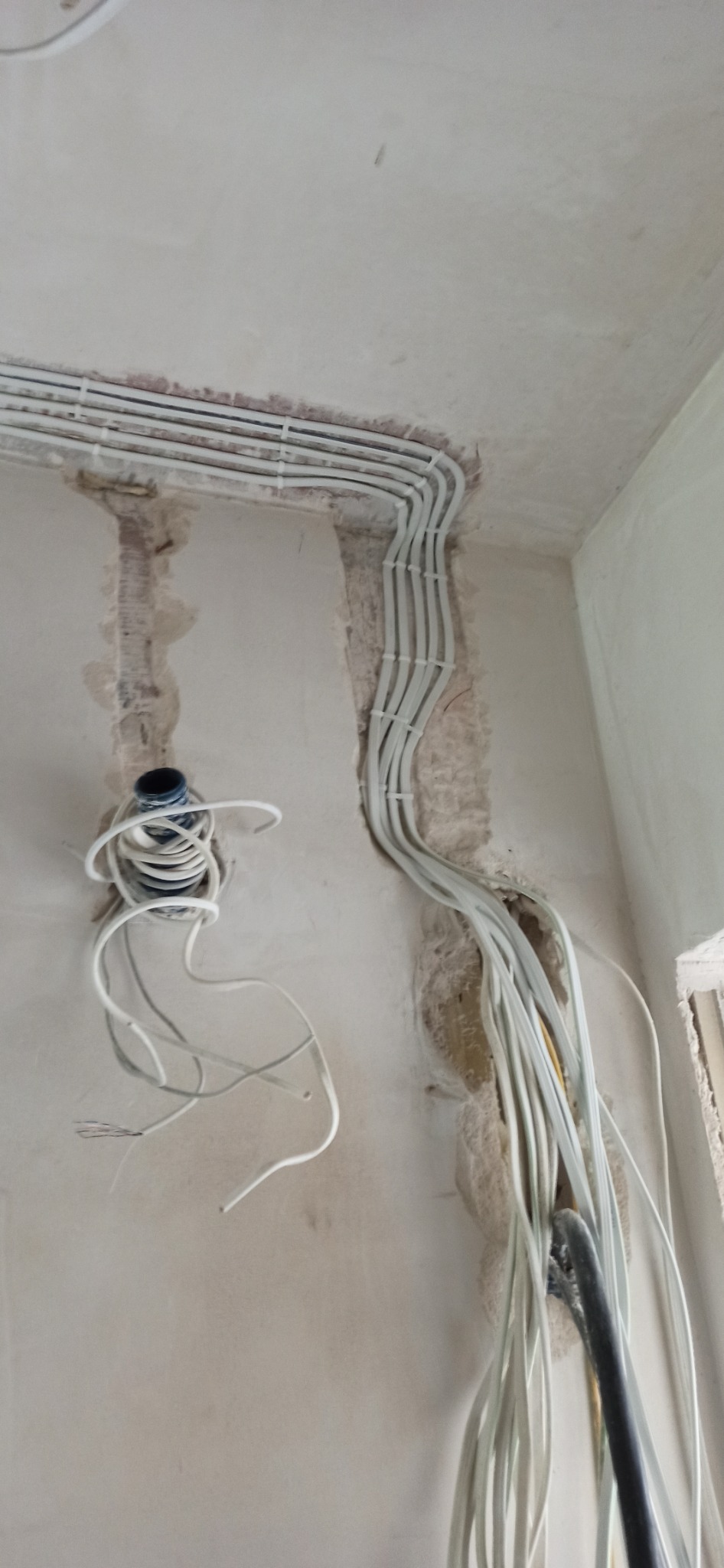 Białe przewody elektryczne ułożone w bruzdach w ścianie, zabezpieczone plastikowymi uchwytami, widoczny otwór z przewodem i puszką instalacyjną.