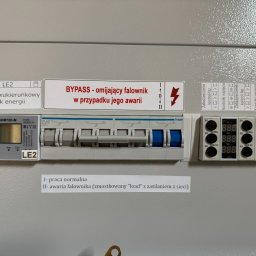 EL-MATIC - Szara szafa elektryczna z zamontowanym dwukierunkowym licznikiem energii, wyłącznikami nadprądowymi Hager z niebieskimi dźwigniami oraz modułami kontroli napięcia, oznaczona naklejkami...