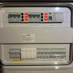 Rozdzielnica elektryczna z zabezpieczeniami, wyłącznikami i modułami pomiarowymi Hager i Schneider. Widoczne oznaczenia: zasilanie z sieci, rezerwowe, falownik, agregat.