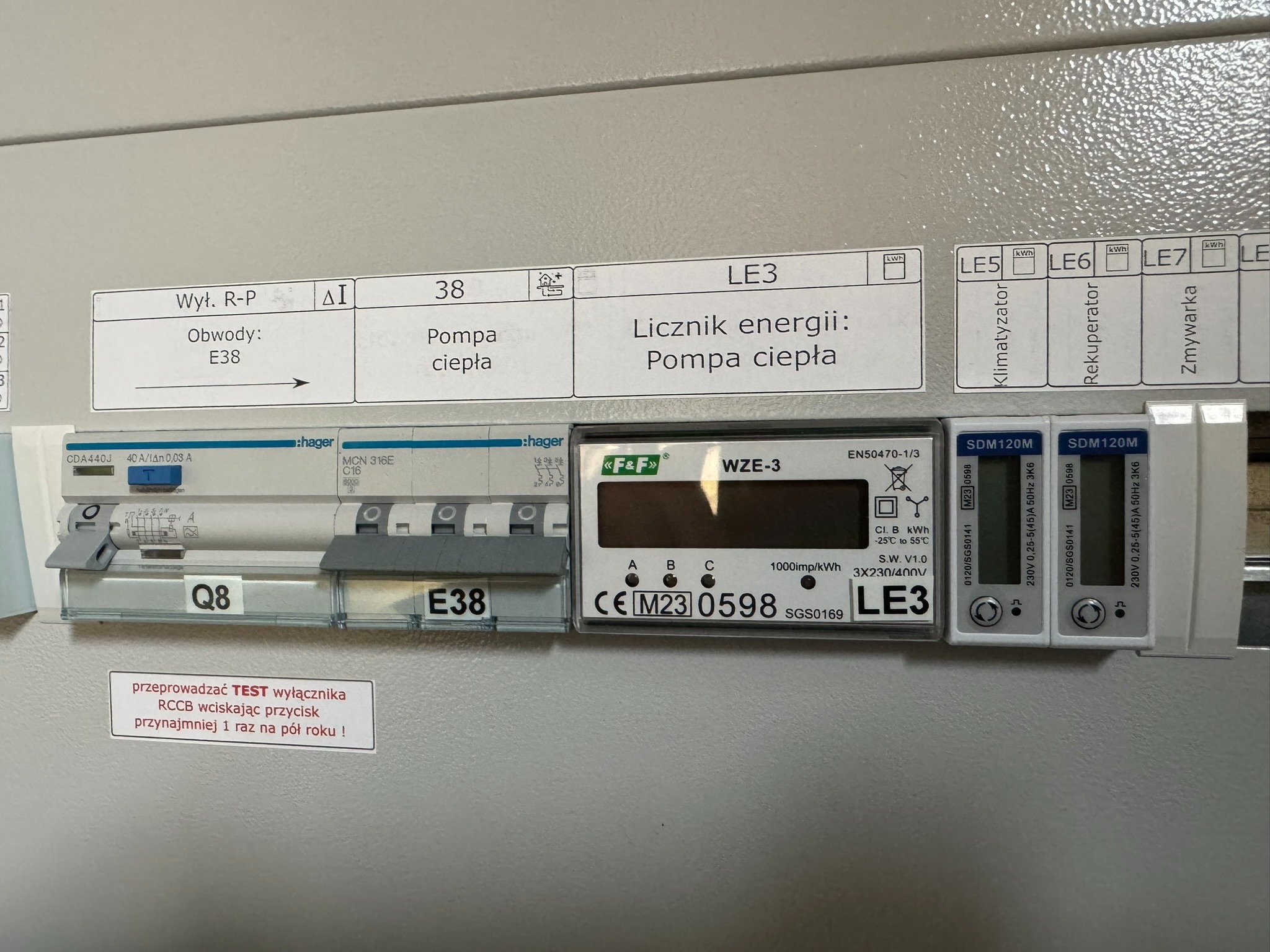 Rozdzielnica elektryczna z zamontowanymi licznikami energii elektrycznej, wyłącznikiem różnicowoprądowym, opisana jako obwód pompy ciepła, z naklejką przypominającą o regularnym teście wyłącznika...